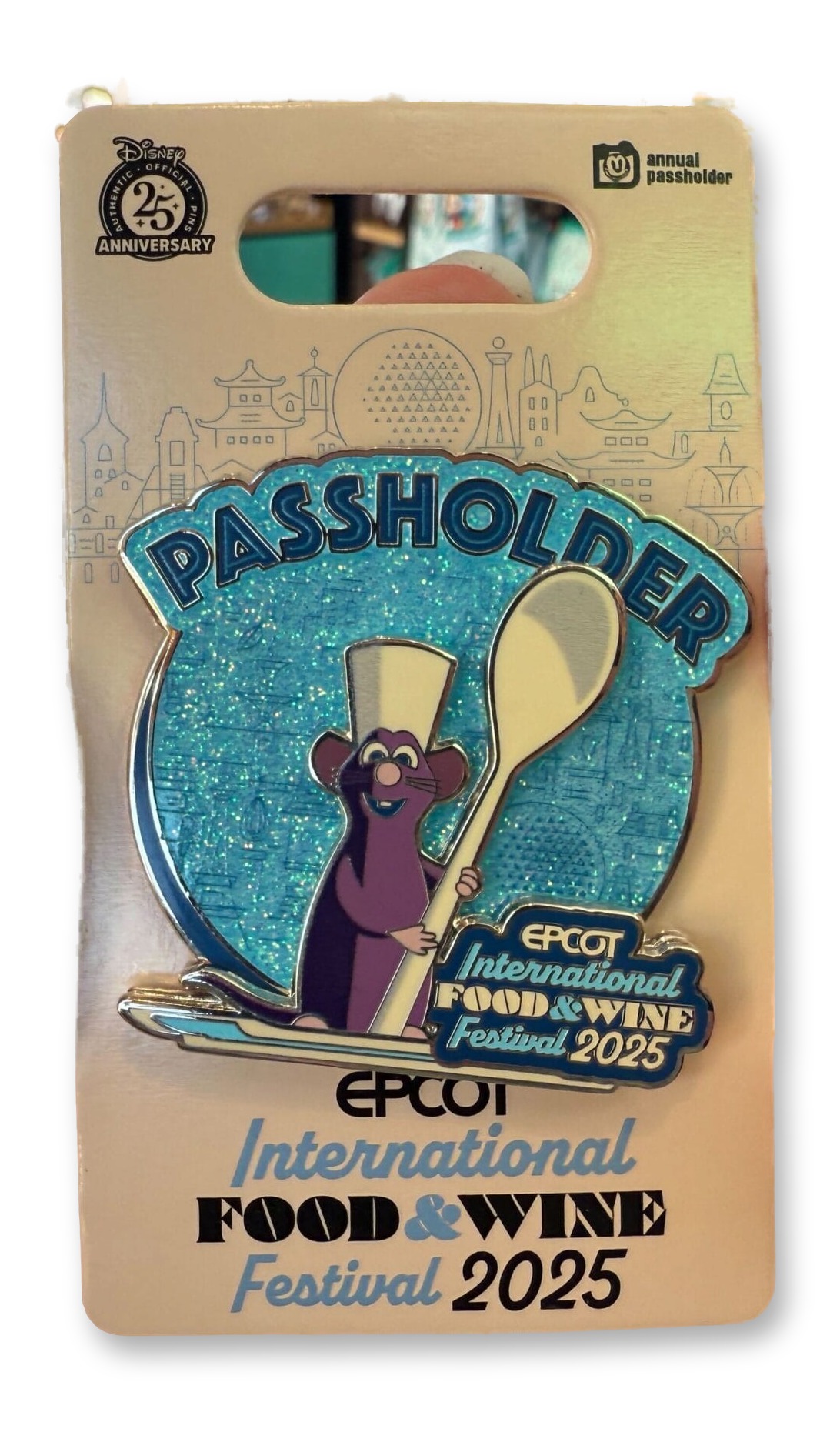 A hand holds a Chef Remy Annual Passholder pin on its retail card inside an EPCOT gift shop; the pin shows Remy in a chef's toque holding a spoon over a glittering teal 'PASSHOLDER' badge and the Food & Wine Festival 2025 logo.