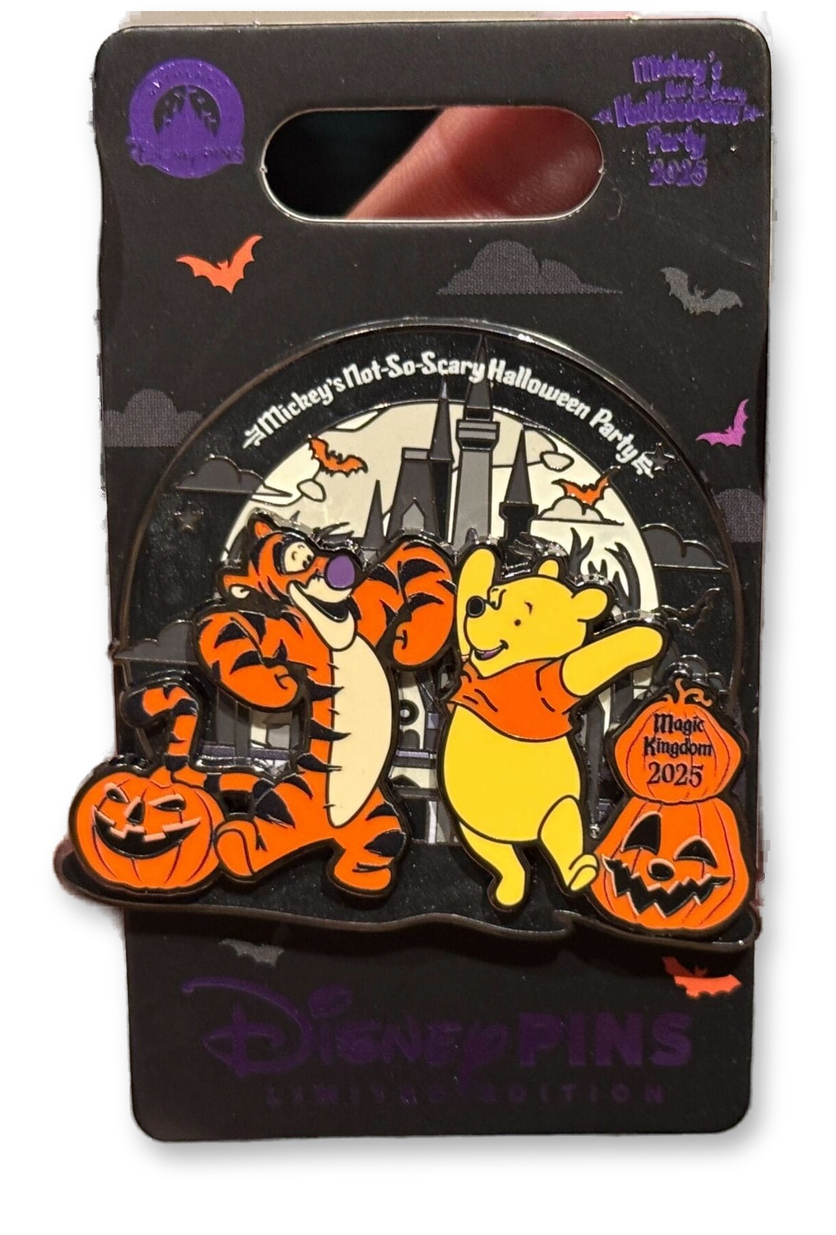 A hand holding an enamel Disney trading pin on its retail card, showing Tigger bouncing beside Winnie the Pooh in front of a black-and-white Cinderella Castle with jack-o'-lanterns, bats, and 'Mickey's Not-So-Scary Halloween Party 2025' text.