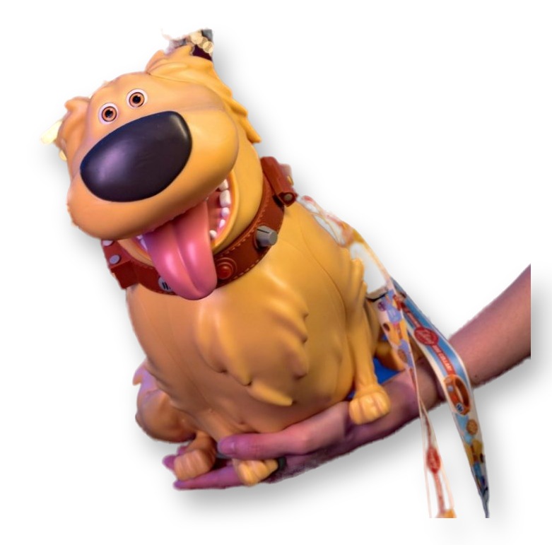 Tan popcorn bucket sculpted as Dug the dog from Up, with a brown collar, floppy tongue, and Magic Key Pixar Fest lanyard, held up in front of the lit Pixar Pier sign at dusk.