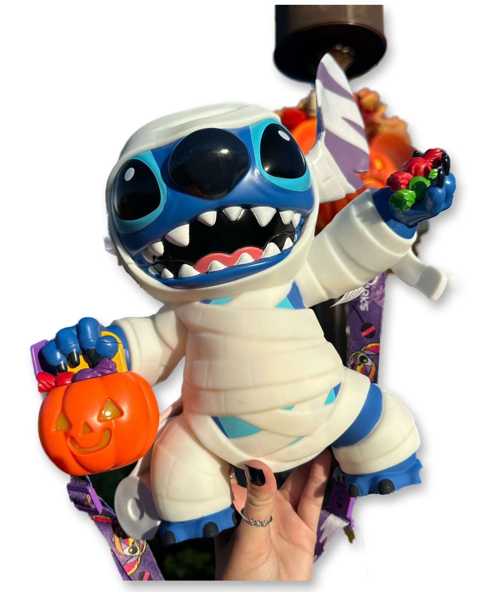 A plastic popcorn bucket shaped like Stitch wrapped in mummy bandages, mouth open baring fangs, holding a glowing jack-o'-lantern candy pail, photographed outdoors near Magic Kingdom's Main Street hub.