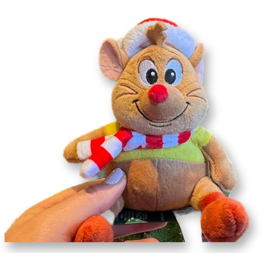 A plush Gus Gus mouse from Cinderella wearing a Santa hat and red-and-white scarf, held up in front of a decorated Christmas tree at a Disney Parks shop.